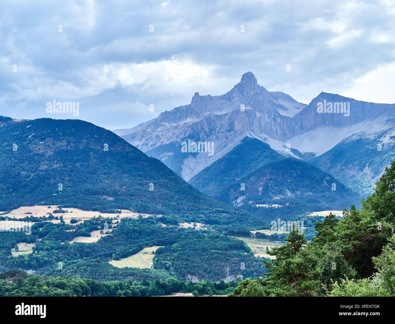 Dramatischer Berggipfel über einer grünen Talloandschaft in den Hautes Alpes in Frankreich, mit bewaldeten Hügeln und bewölktem Himmel Stockfoto