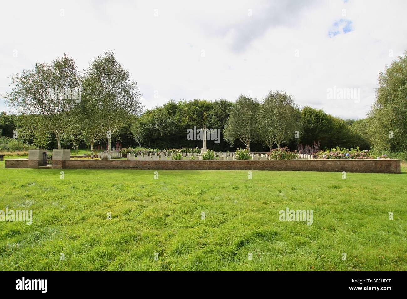 Das weiße steinerne Kreuz der Opferung steht zwischen Reihen von Grabsteinen hinter einer niedrigen Backsteinmauer auf einem Commonwealth-Militärfriedhof in Belgien. Stockfoto