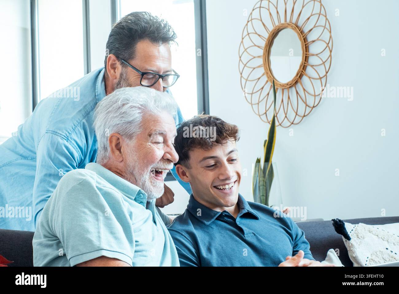 Eine multigenerationale Familie teilt einen freudigen Moment auf einem Wohnzimmersofa, während ein Großvater, ein Vater und ein jugendlicher Enkel gemeinsam lachen und Wärme zeigen Stockfoto