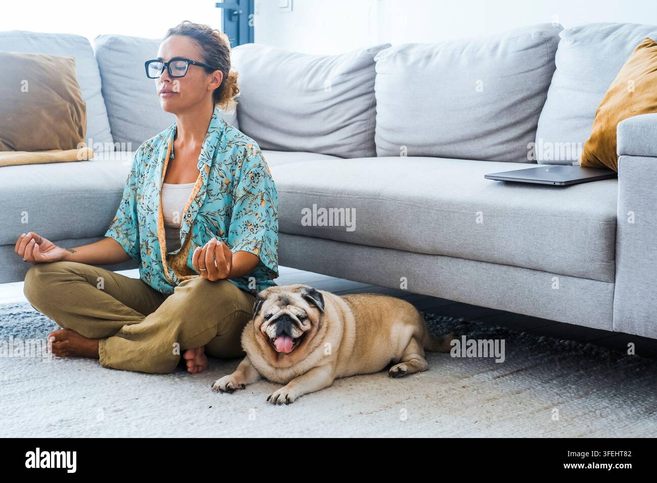 Frau meditiert auf einem Teppich mit ihrem Mops neben ihr in einem hellen Wohnzimmer, praktiziert Achtsamkeit und Entspannung zu Hause mit einem Laptop auf der Couch. Stockfoto