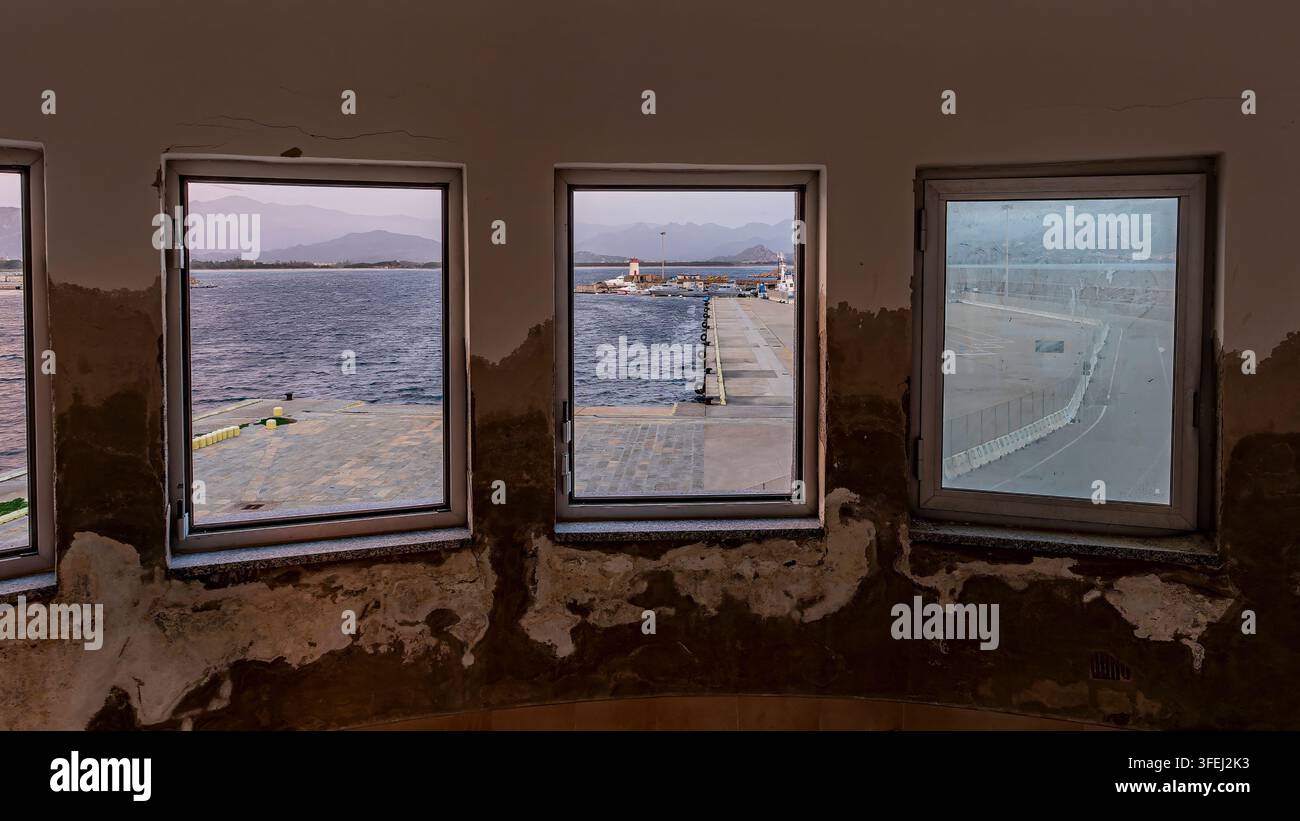 Verlassenes Gebäudeinterieur mit abblätternder Farbe, das durch mehrere Fenster Ausblicke auf den Hafen von Tortoli, den Pier und die Berge bietet Stockfoto