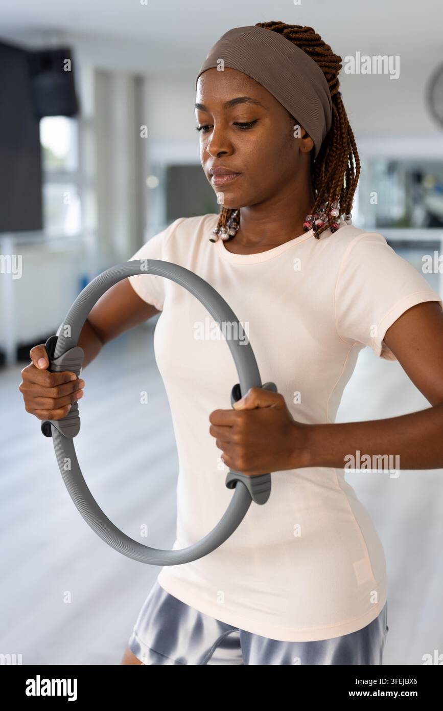 Afroamerikanische Frau drückt grauen Pilatesring, trägt Stirnband und enganliegendes Oberteil im Studio Stockfoto