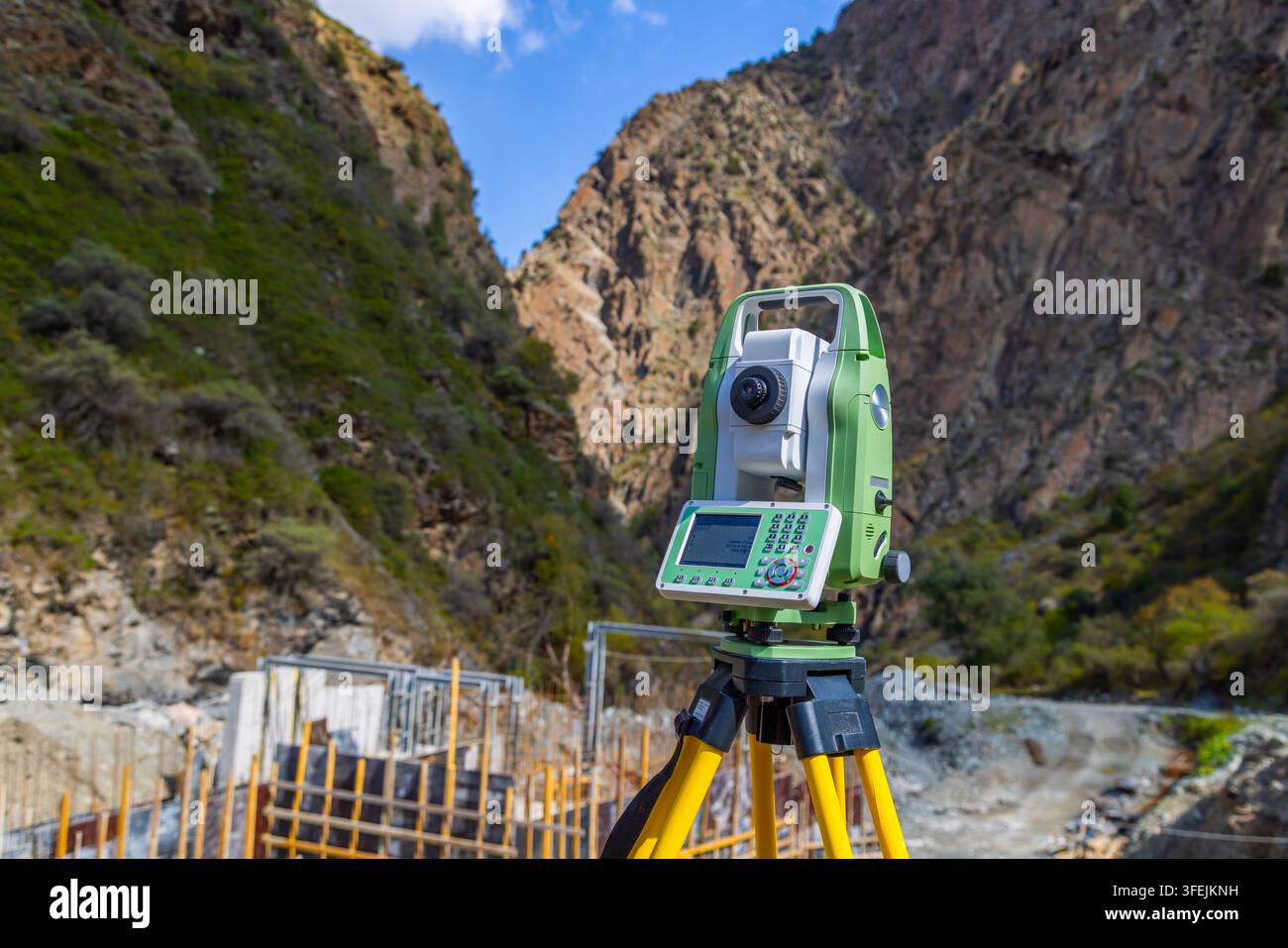 Ein grün-weißes Totalstation-Vermessungsinstrument ist auf einem gelben Stativ in einem rauen bergigen Gebiet in Kirgisistan montiert. Das Digitaldisplay zeigt Daten an, wobei im Hintergrund eine Baustelle zu sehen ist, die laufende Infrastrukturarbeiten anzeigt. Stockfoto