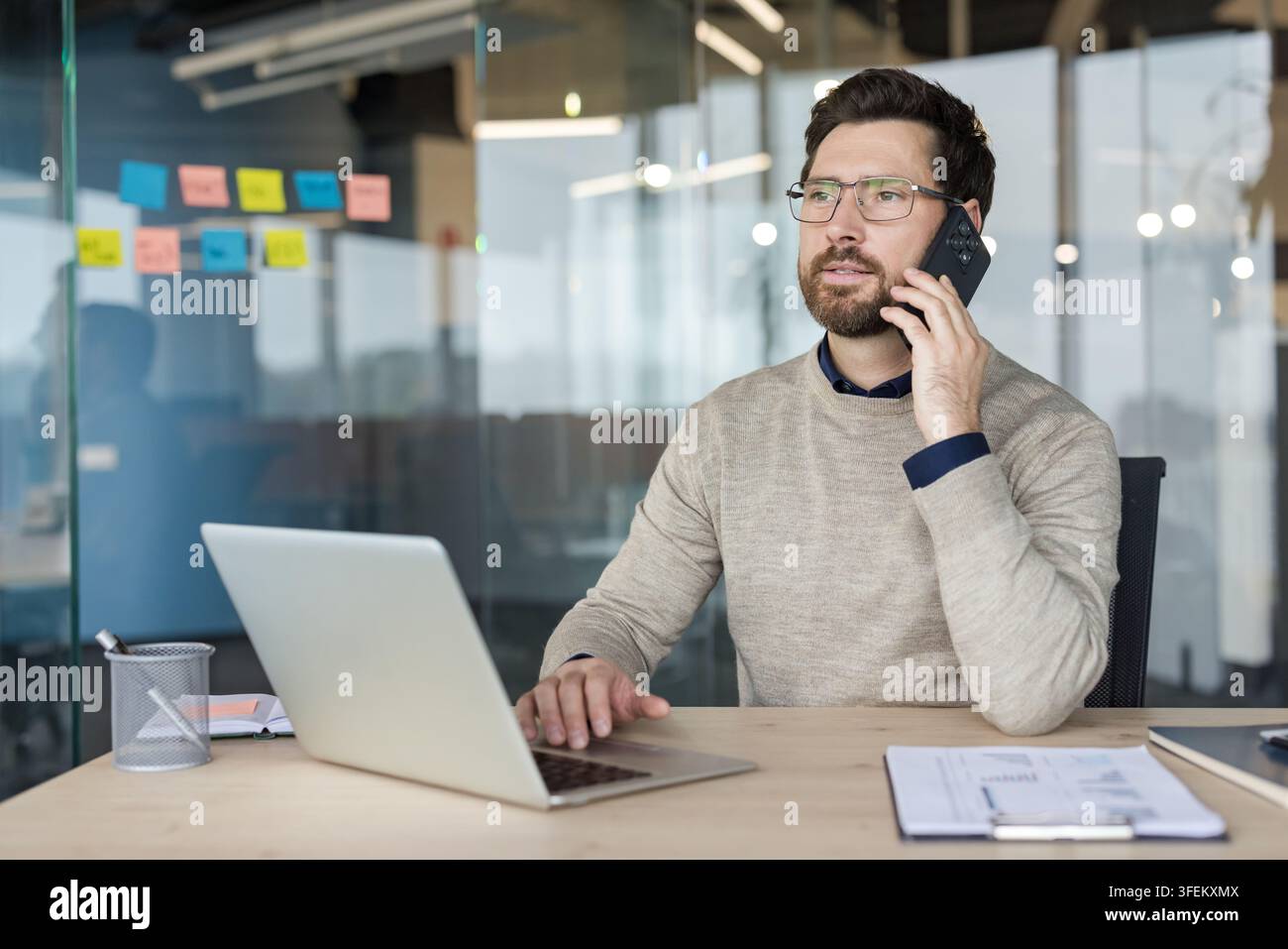 Professioneller Mann führt ein Geschäftsgespräch am Mobiltelefon, nutzt einen Laptop an seinem Schreibtisch in einem modernen Büro, symbolisiert Fernarbeit, Kommunikation und Konnektivität Stockfoto