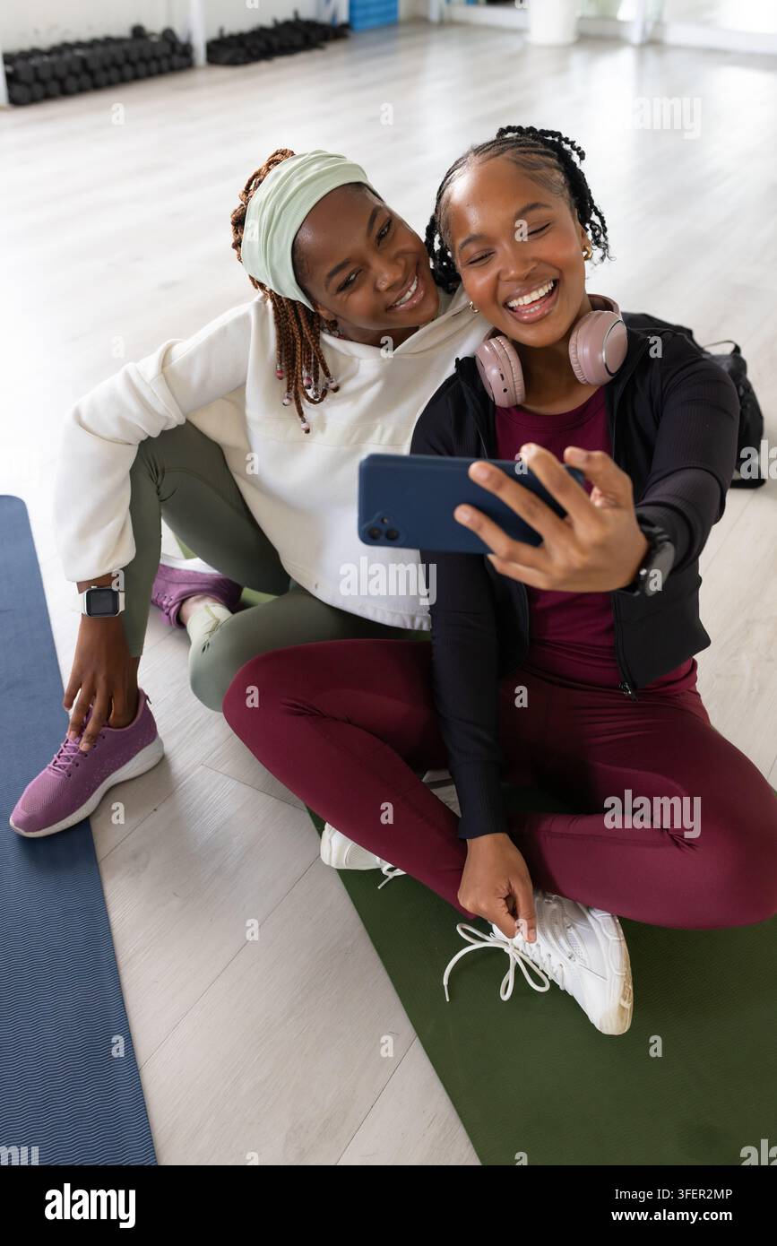 Zwei afroamerikanische Frauen sitzen auf Yogamatten und machen ein Smartphone-Selfie in einem hellen Fitnessstudio Stockfoto