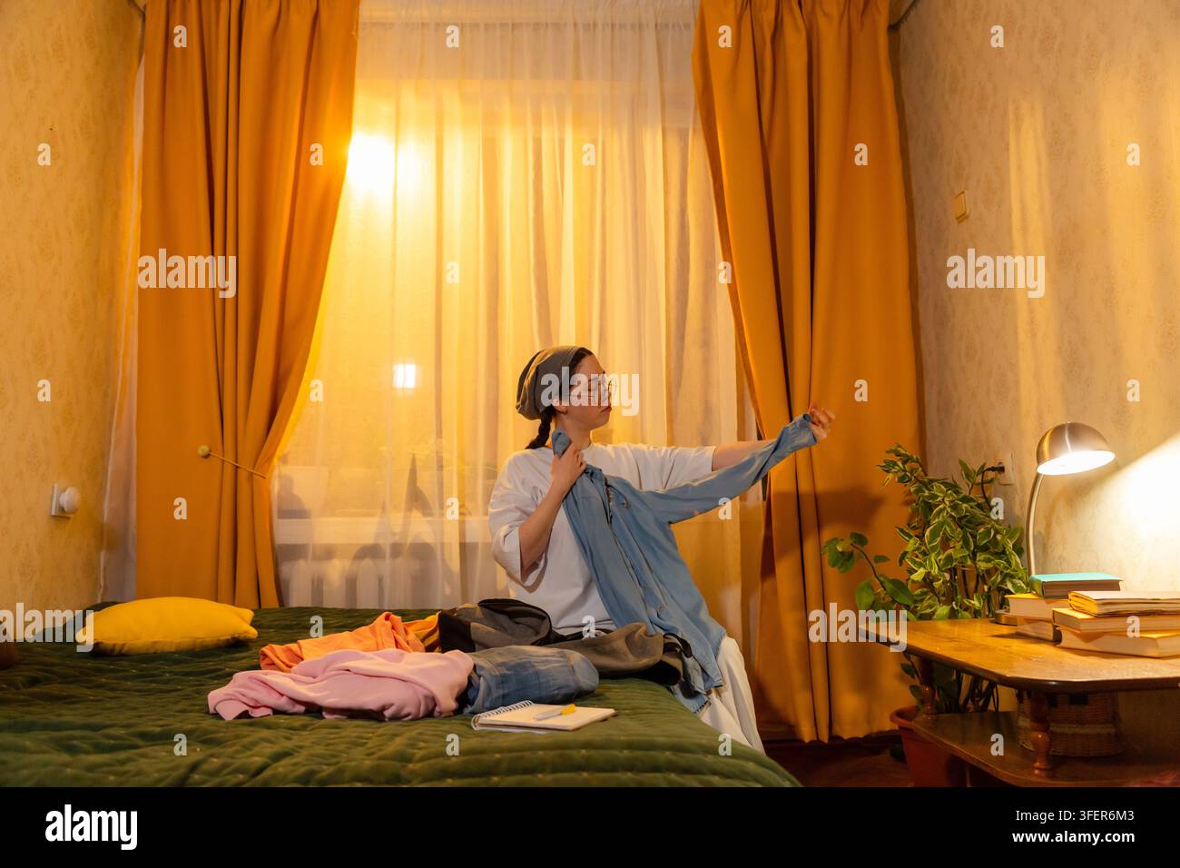 Kaukasische Frau faltet Wäsche in ihrem Zimmer, während die Sonne untergeht. Sie ordnet die Gegenstände sorgfältig an. Konzept der Raumorganisation. Niedriger Winkel, Weitwinkelaufnahme. Stockfoto