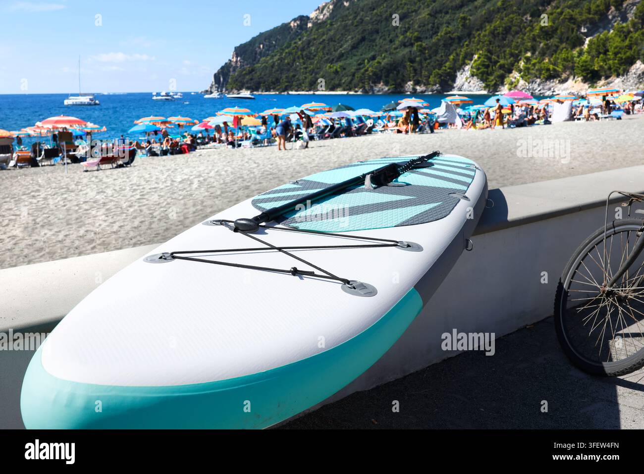 Aufblasbares Stand Up Paddelboard mit Paddel, das an einer Strandmauer lehnt. Aufblasbares SUP-Board an einem sonnigen Sommertag in einem überfüllten Strandresort am Meer. Aktives Wasser Stockfoto