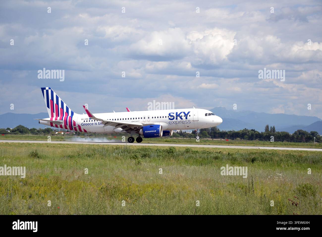 SKY express Airbus A320neo-Passagierflugzeug landet auf der Landebahn mit Reifenrauch unter bewölktem Himmel, Berge im Hintergrund. Stockfoto