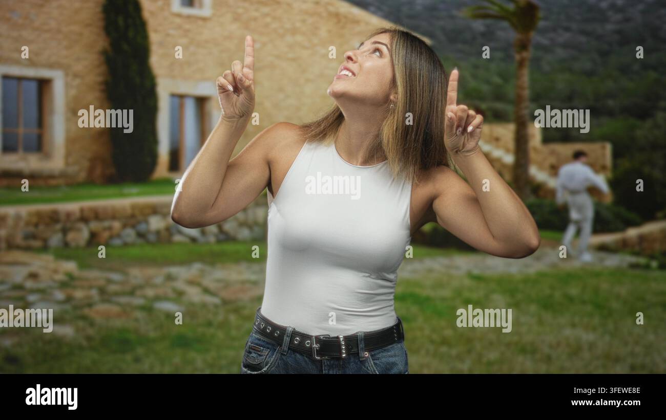 Frau zeigt mit sichtbaren Händen die Zeigefinger nach oben und lächelt vor dem Steingebäude, trägt weiße Tanktops und Jeans; verspielte Freude. Stockfoto