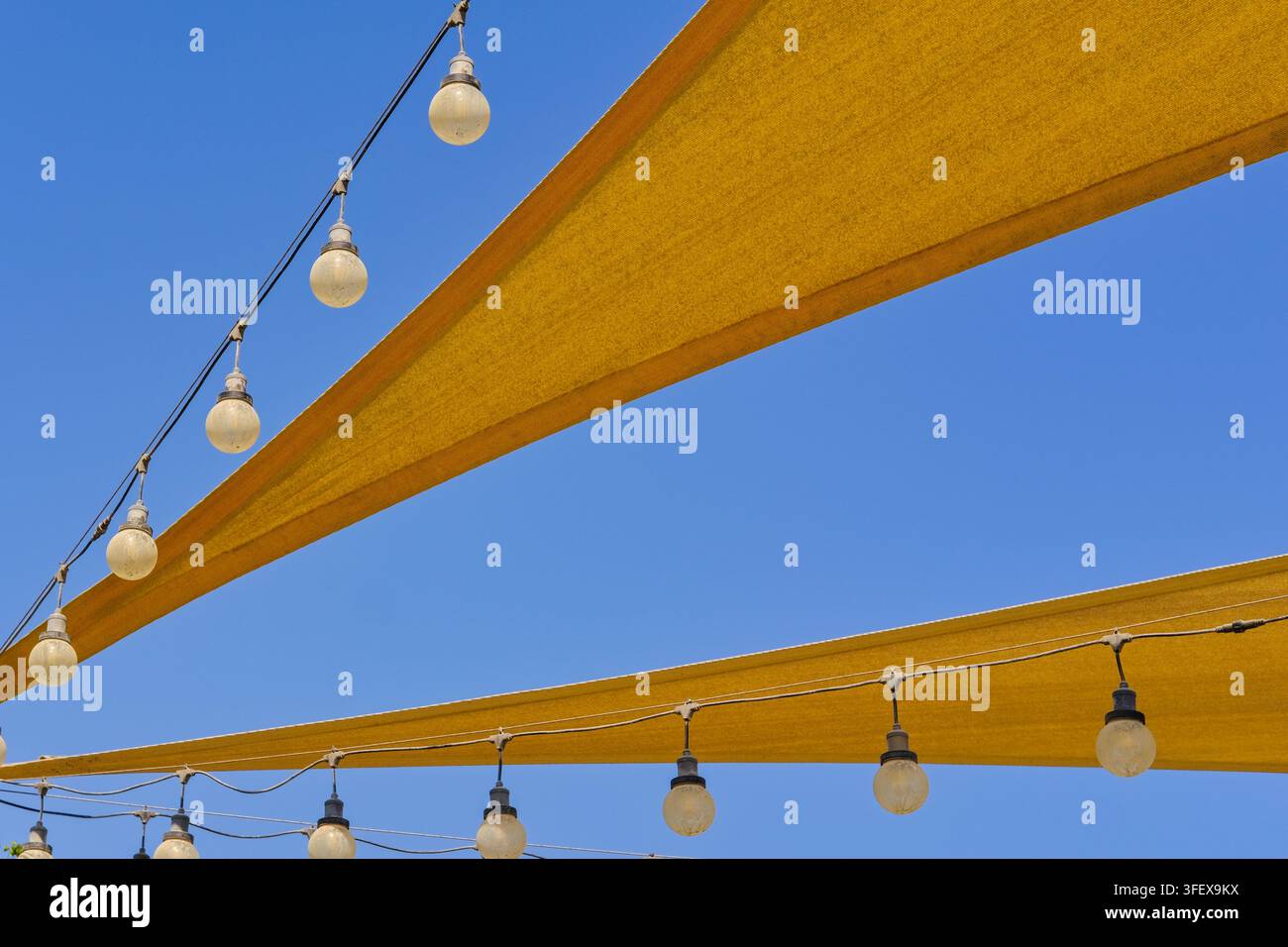 Gespannte Leinwand-Sonnensegel und hängende Glühbirnen-Lichter bilden einen dynamischen geometrischen Hintergrund vor blauem Himmel und kombinieren Sonnenschutz und Außenbeleuchtung Stockfoto