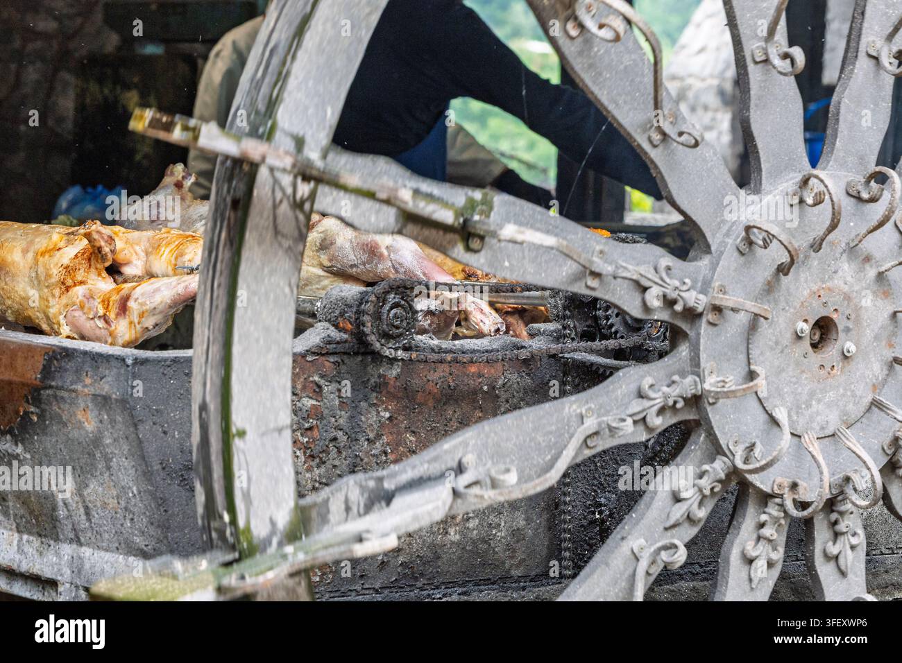 Ganze Lämmer drehen sich langsam an einem Metallspieß hinter einem verzierten Wagenrad, über Holzkohle in einer rustikalen Außenumgebung geröstet, und rufen traditionelle Straßen... Stockfoto