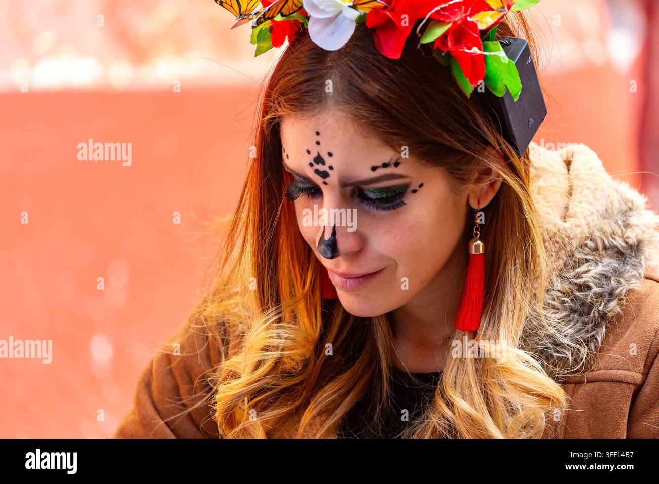 SAN MIGUEL DE ALLENDE, MEXIKO – 01. NOVEMBER 2019: Eine Frau mit Schmetterlings- und Blumenkopfschmuck und Gesichtsfarbe schaut nach unten. Sie trägt einen braunen Mantel w Stockfoto