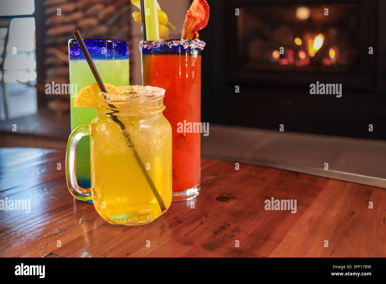 Drei bunte Mixgetränke mit Zitrus-, Sellerie- und Speck-Garnierungen stehen auf einem Holztisch neben einem leuchtenden Kamin im Innenraum. Stockfoto
