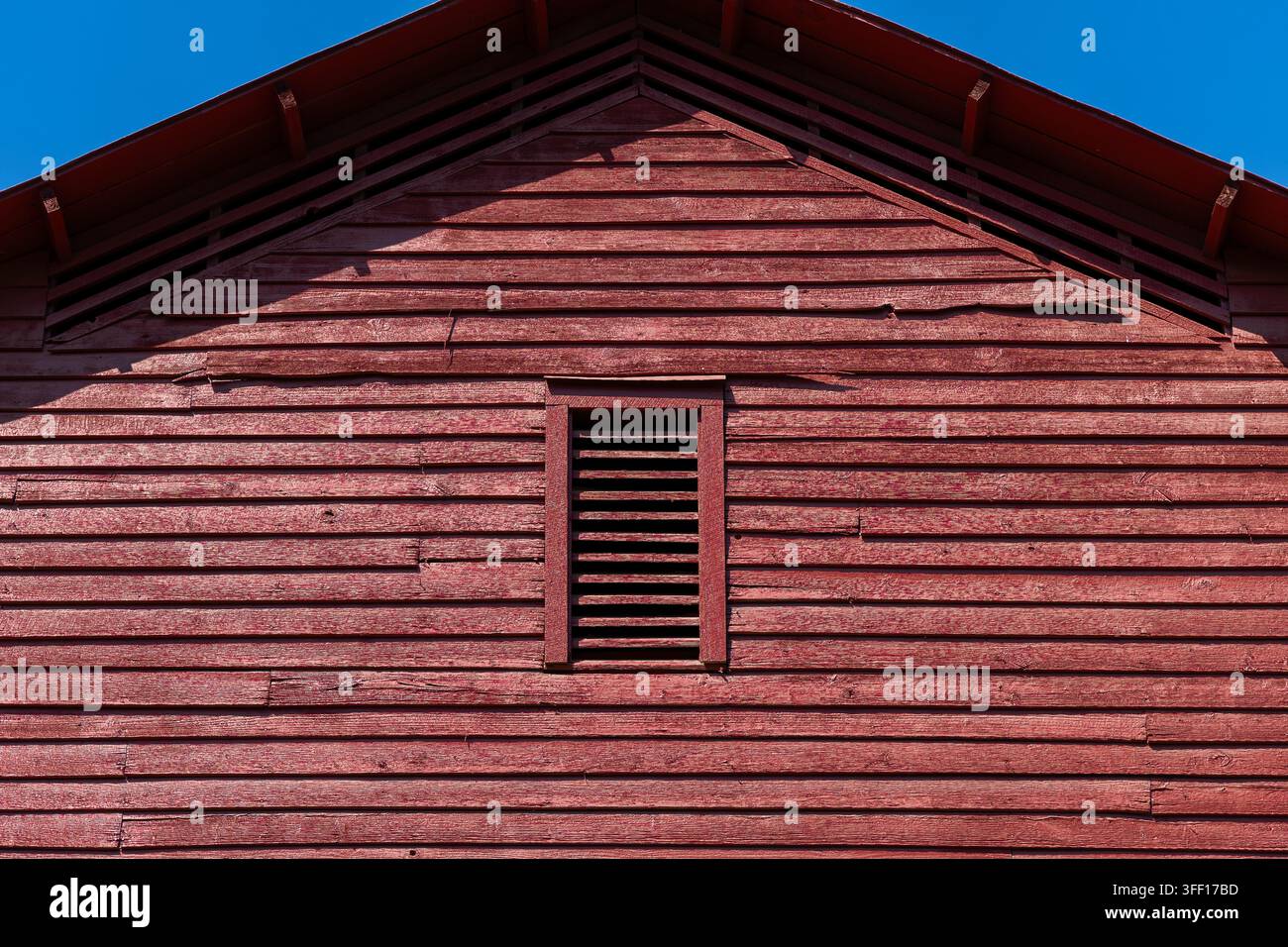 xGeschnittene architektonische Ansicht des Giebels eines verwitterten roten Holzbarn mit horizontaler Schindeln, zentraler Lüftungsöffnung mit Lamellen und tiefen geometrischen Schatten b Stockfoto