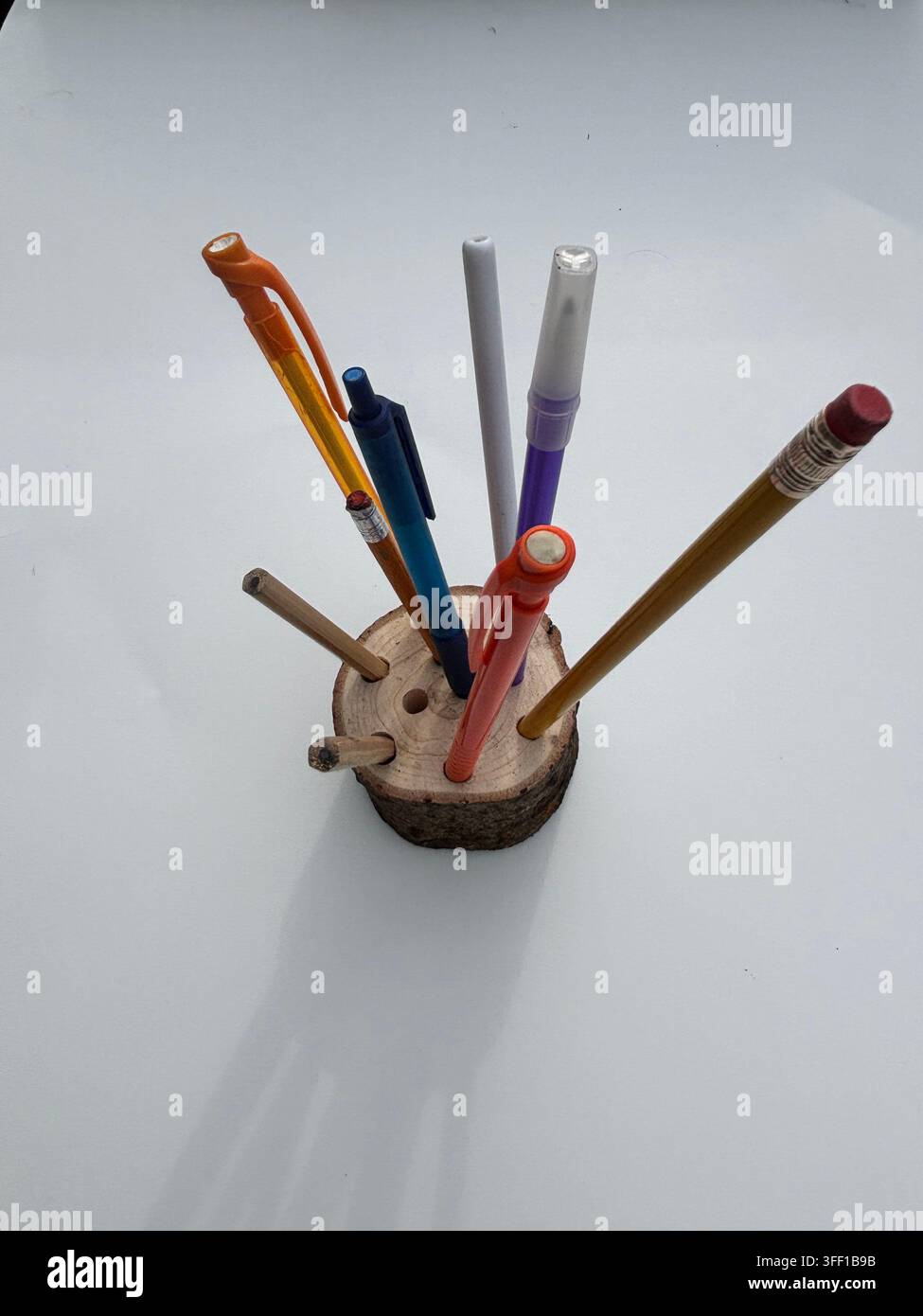 Ein rustikaler, handgefertigter Stablampenhalter aus Holz, gefüllt mit einer Auswahl an farbenfrohen Stiften, Bleistiften und Markern auf einem sauberen weißen Hintergrund - Smartphone-aufgenommenes Stockfoto