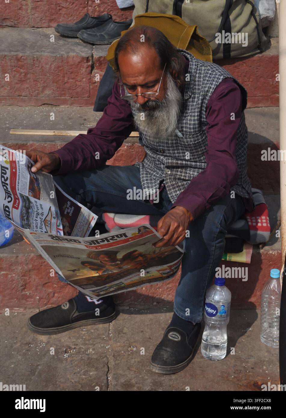 VARANASI, INDIEN - 22. JANUAR 2026: Ein älterer bärtiger Mann sitzt auf den Ghat-Stufen und liest eine Zeitung auf Hindi. Stockfoto