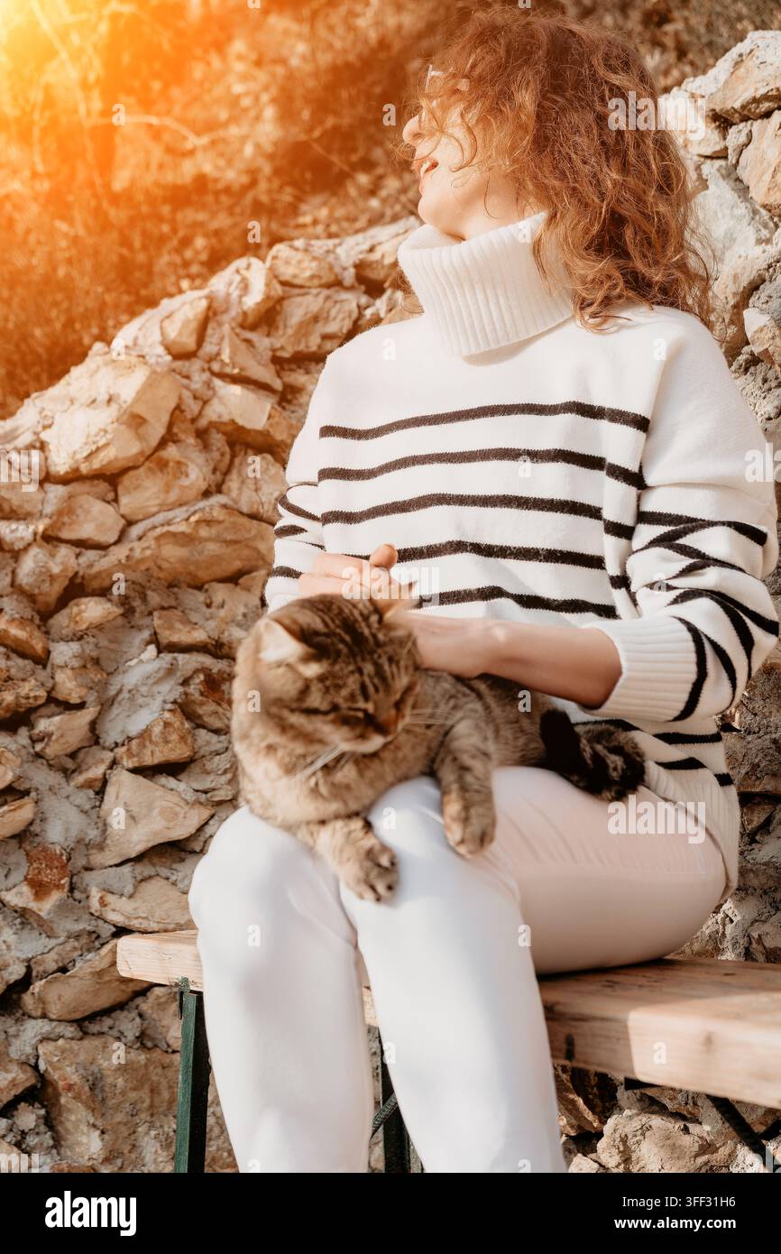 Frau, die eine junge Frau in gestreiftem Pullover streichelt, sitzt mit getigerter Katze im goldenen Sonnenlicht an einer Steinmauer auf einer Bank Stockfoto