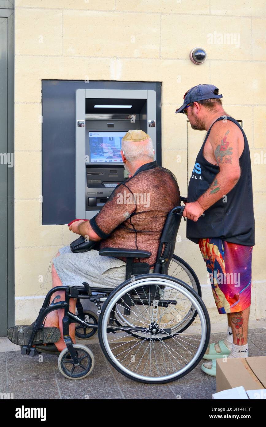Behinderter Mann mittleren Alters im Rollstuhl mit Helfer in jazzigen Shorts, der Geld am Bankgeldautomaten abhebt – Loches, Indre-et-Loire (37), Frankreich. Stockfoto