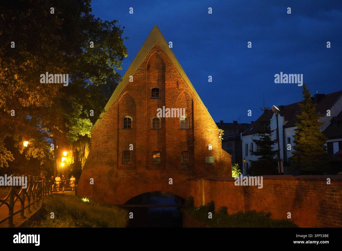 Beleuchtetes mittelalterliches Großmühlengebäude in Danzig, Polen bei Nacht in der Nähe des Kanals mit sanfter Abendbeleuchtung und Bäumen. Stockfoto