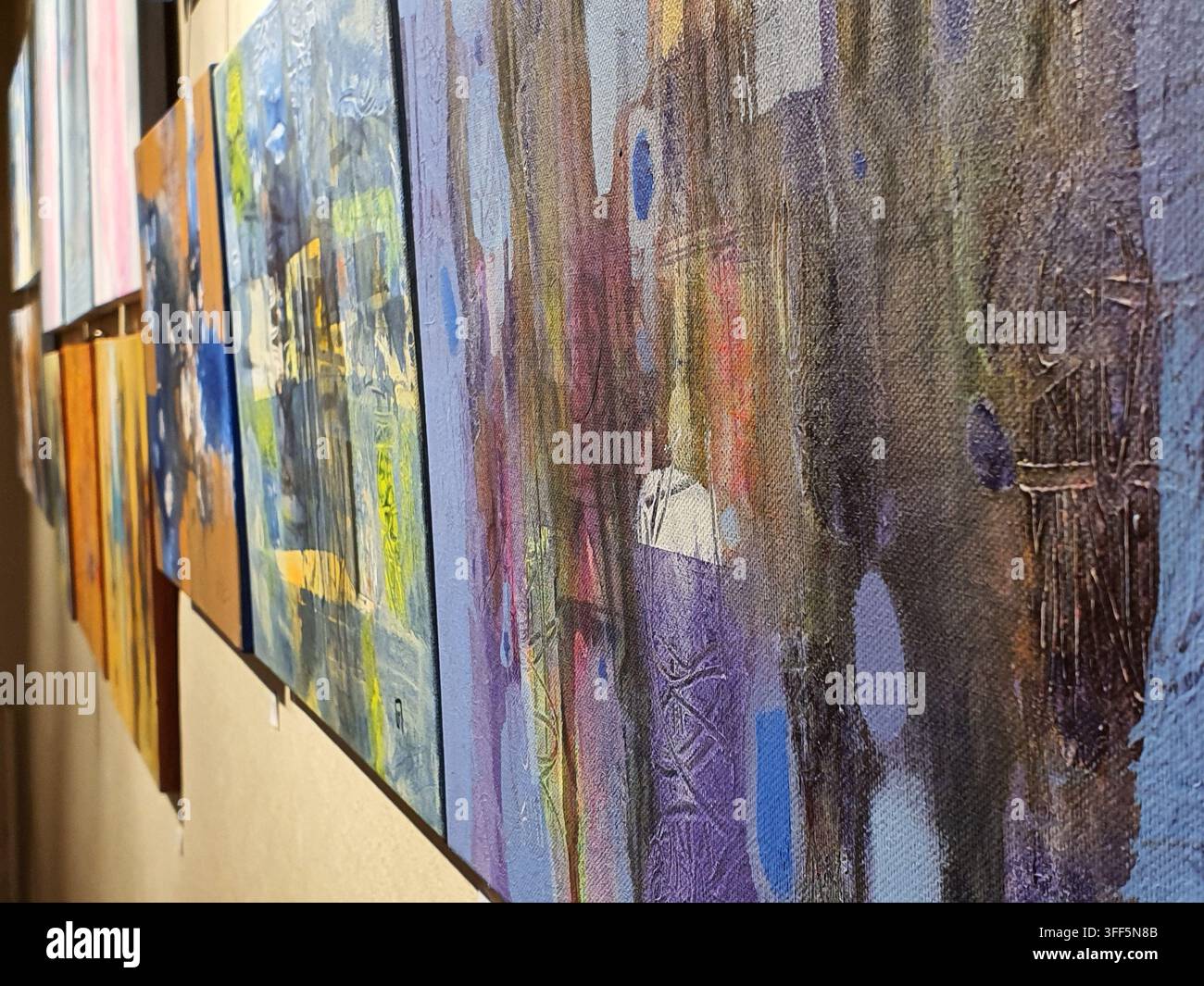 Eine Nahaufnahme, seitliche Ansicht von mehreren abstrakten Gemälden, die in einer Reihe an einer hellen Wand hängen. Die Leinwände zeigen verschiedene Farben, darunter Stockfoto