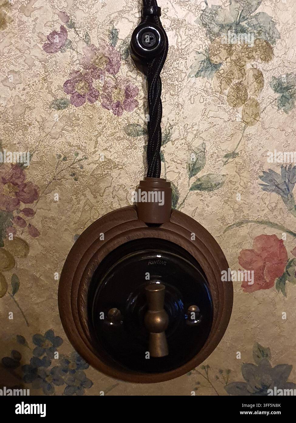 Eine Nahaufnahme eines elektrischen Lichtschalters im Vintage-Stil, der auf floraler Tapete montiert ist. Der Schalter verfügt über einen runden braunen Bakelit- oder Holzknopf Stockfoto