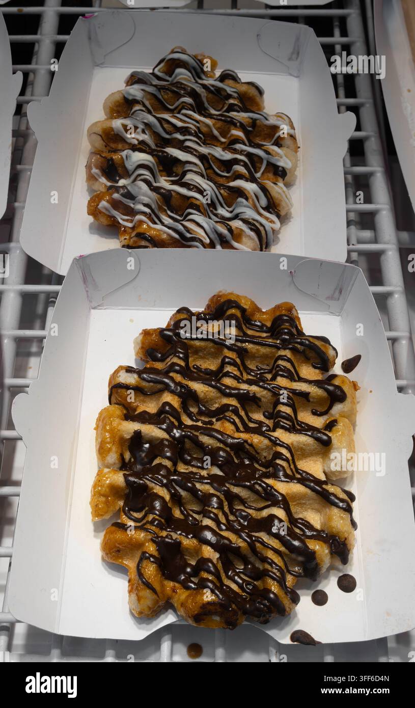 Zwei offene weiße To-Go-Boxen enthalten goldene gebratene belgische Waffel-Snacks, die mit dunkler Schokoladensoße und cremiger weißer Sauce auf einem Metallgestell garniert sind. Stockfoto