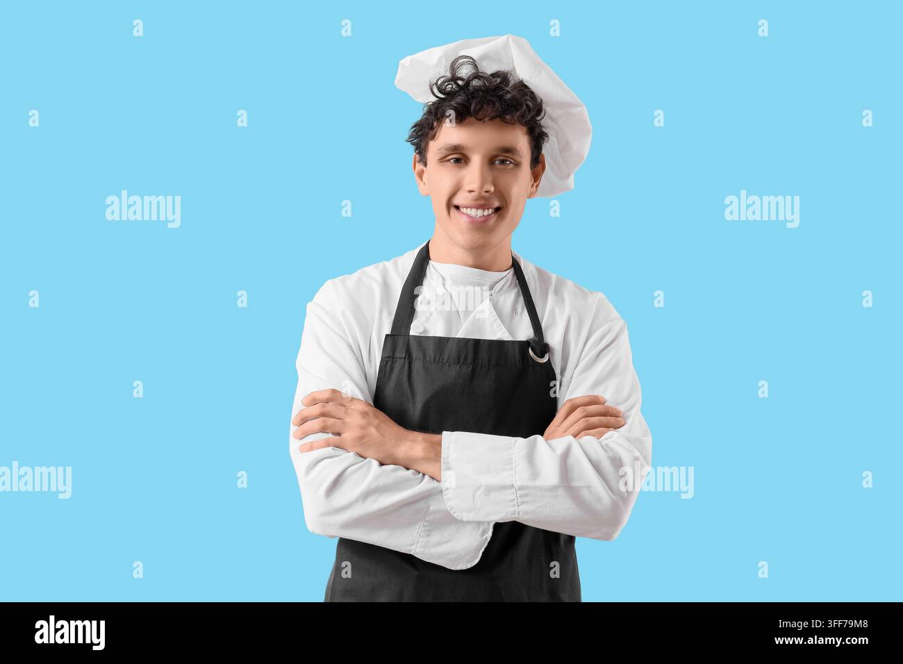 Gastronomieberuf und Hotellerie-Karriere, lächelnder junger Koch in weißer Uniform und schwarzer Schürze, der mit verschränkten Armen auf blauem Hintergrund steht Stockfoto