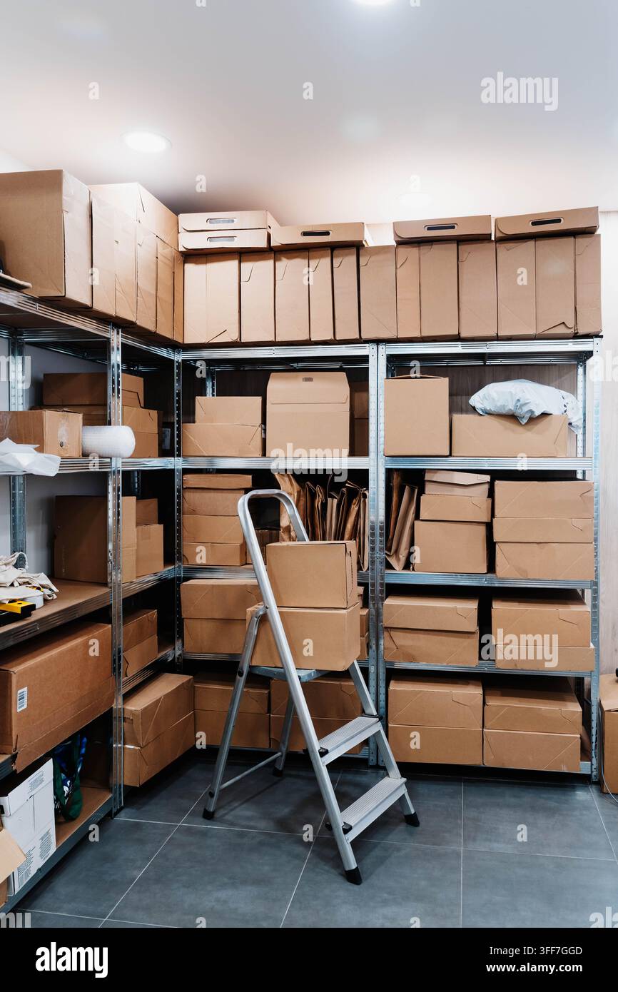 Kartons und Verpackungsmaterial füllen Metallregale in einem kleinen Lagerraum mit einer schiefen Trittleiter. Kompakter Erfüllungsraum für kleine Stockfoto