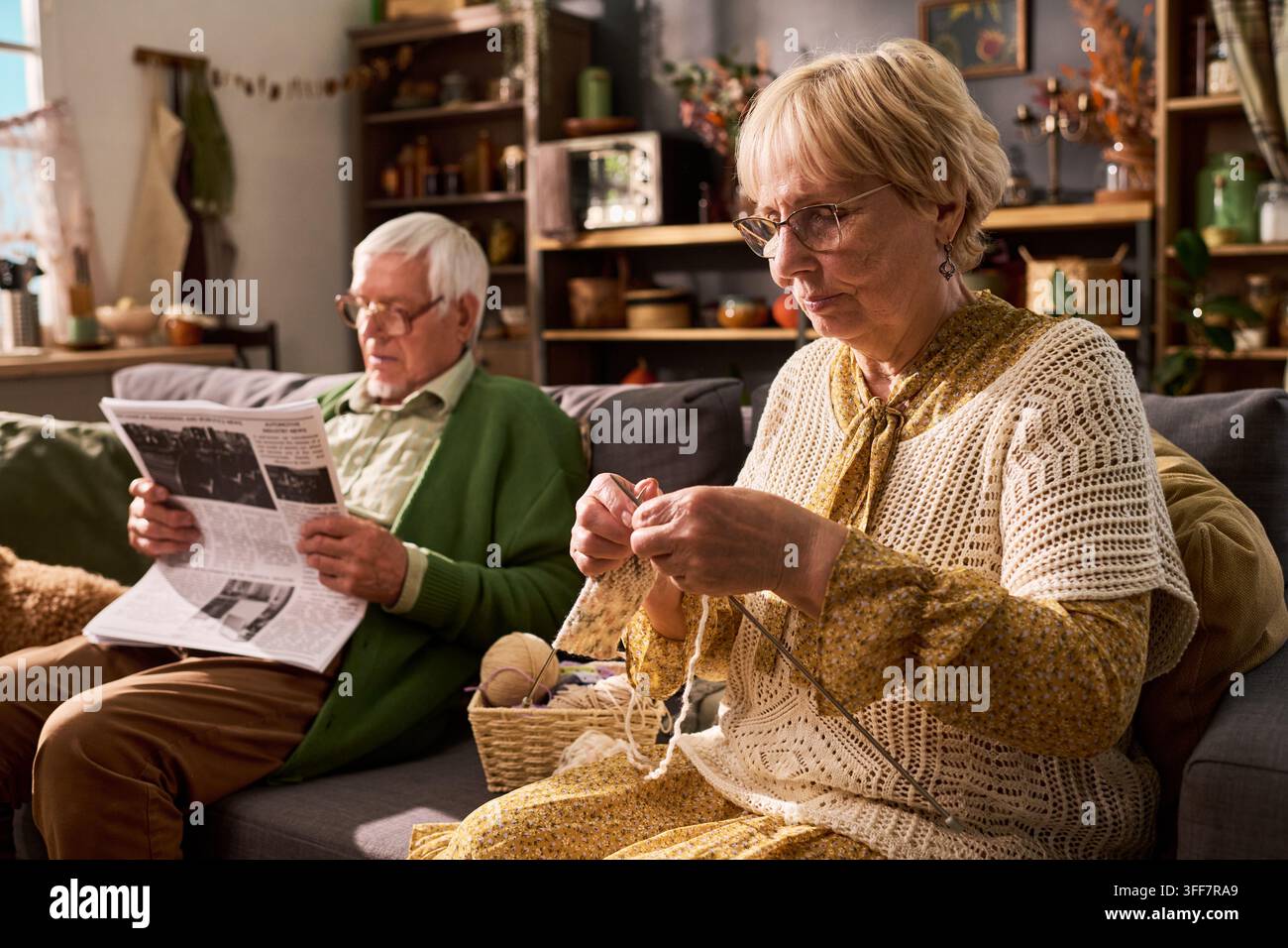 Seniorin strickt neben einem Senior, der Zeitung zu Hause liest Stockfoto