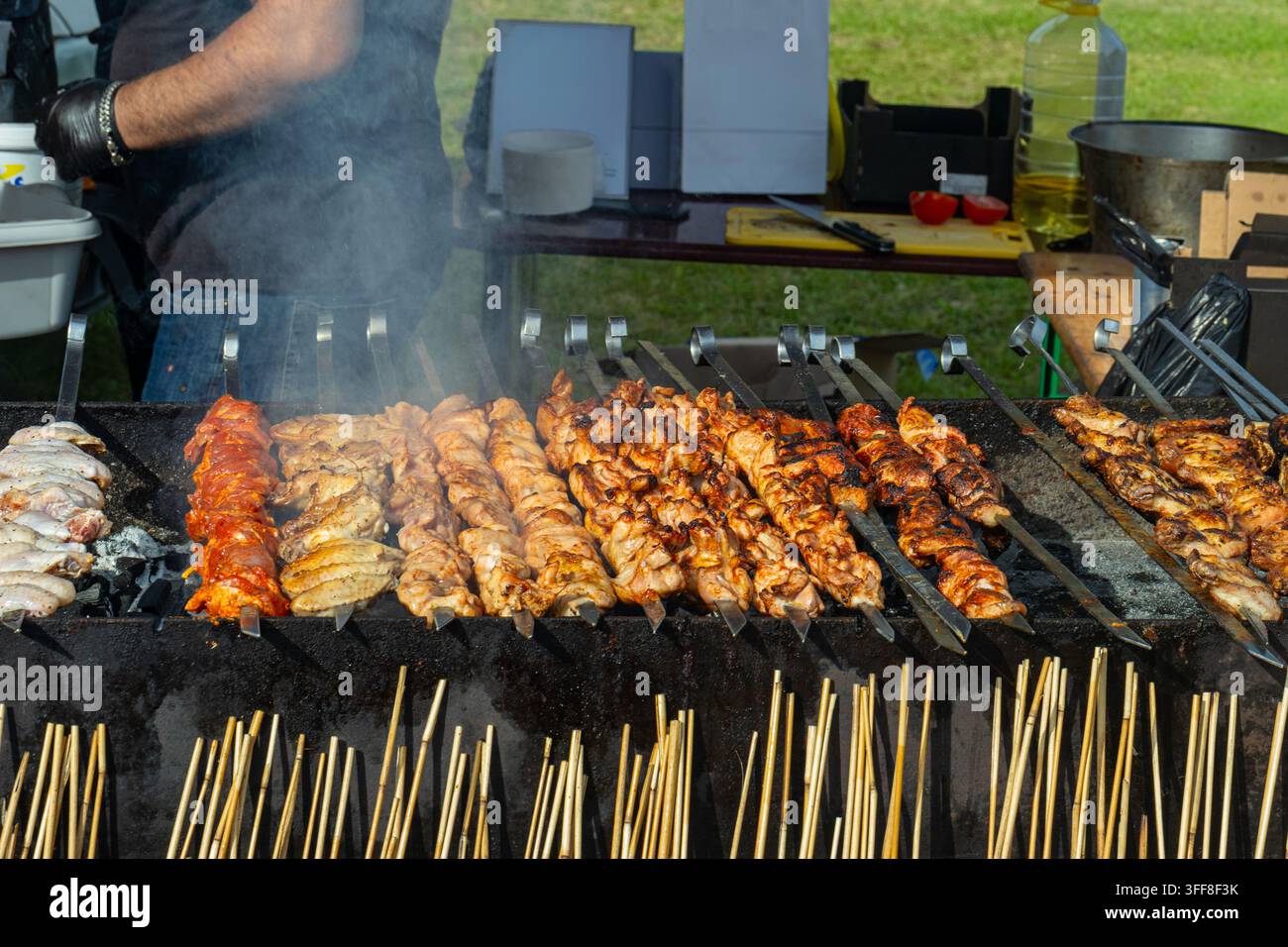 Mittelaufnahme von zahlreichen Metallspießen, beladen mit Hühnerschischkebab, mariniertem Fleisch und Flügeln, die über glühenden Kohlen auf einem großen Außengrill braten Stockfoto