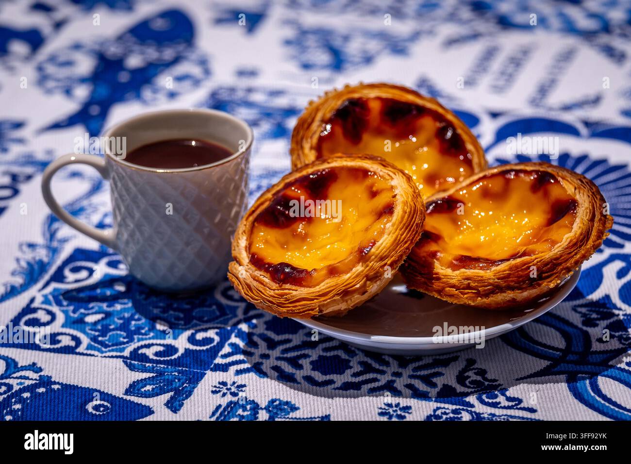 Dreiviertelaufnahme von drei Pastel de Nata-Krustentarten auf weißem Teller und einer kleinen Porzellan-Kaffeetasse mit geometrischem Muster, gefüllt mit frisch gebrühtem Espresso, im Freien auf einer blau-weißen Tischdecke mit traditionellen portugiesischen Azulejo-Fliesenmotiven Stockfoto