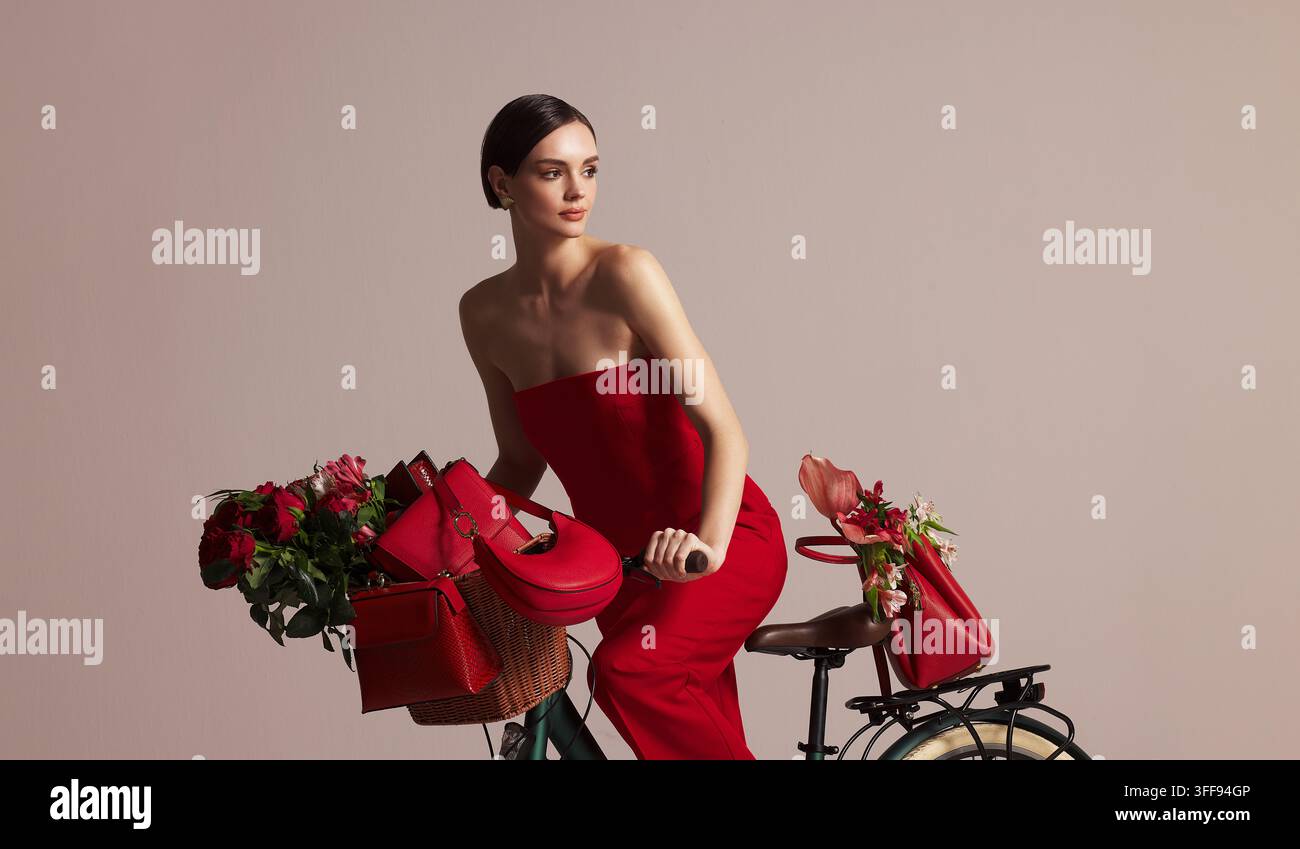Eine hinreißende junge brünette Frau in einem roten Hosenanzug blickt in die Kamera, während sie neben einem Retro-Fahrrad posiert, das mit roten Rosen und Blumen geschmückt ist. Stockfoto