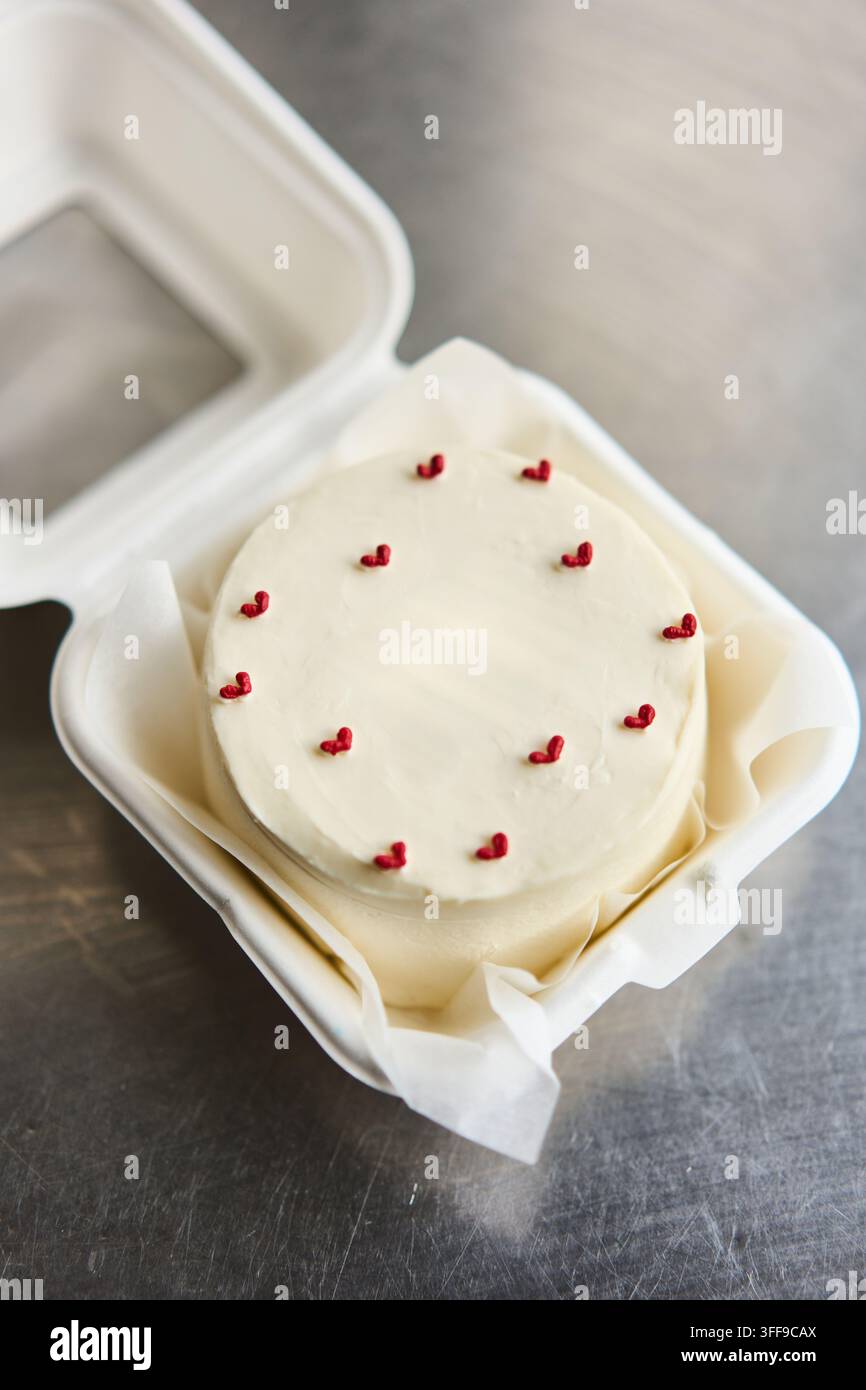 Kleine weiß gefrostete Torte verziert mit winzigen roten Herzstreuseln in einer Mitnahmeschachtel auf Metalloberfläche, mit Torte, Glasur, Weiß, Streusel-Detail, zeigt Stockfoto