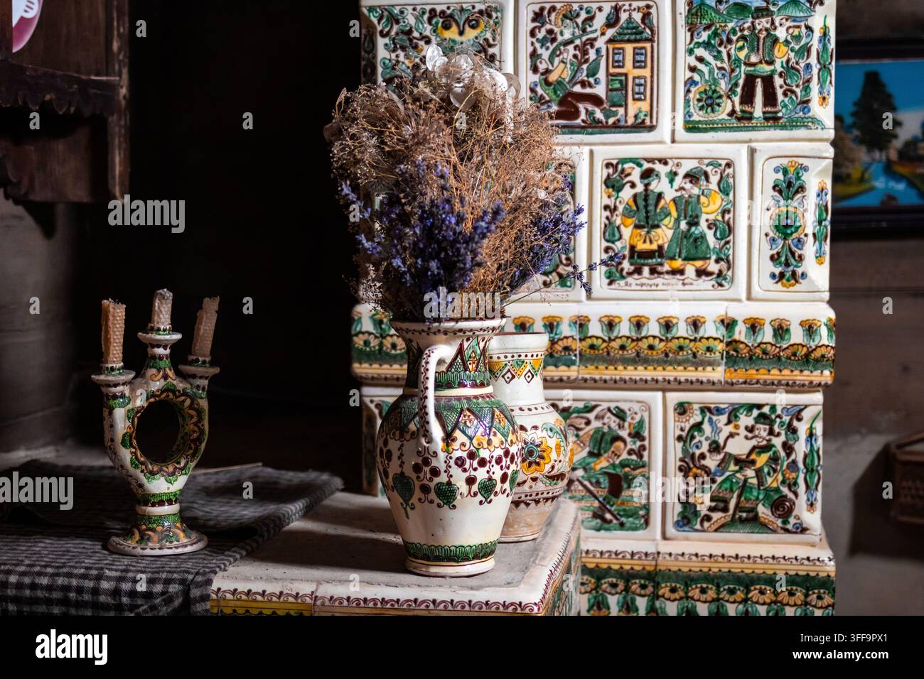 Volkskeramischer Ofen steht neben einem bemalten Krug, der mit getrockneten Blumen gefüllt ist, die Szene ist sanft beleuchtet, um gemusterte Fliesen und handwerkliche Details zu zeigen, die Stockfoto