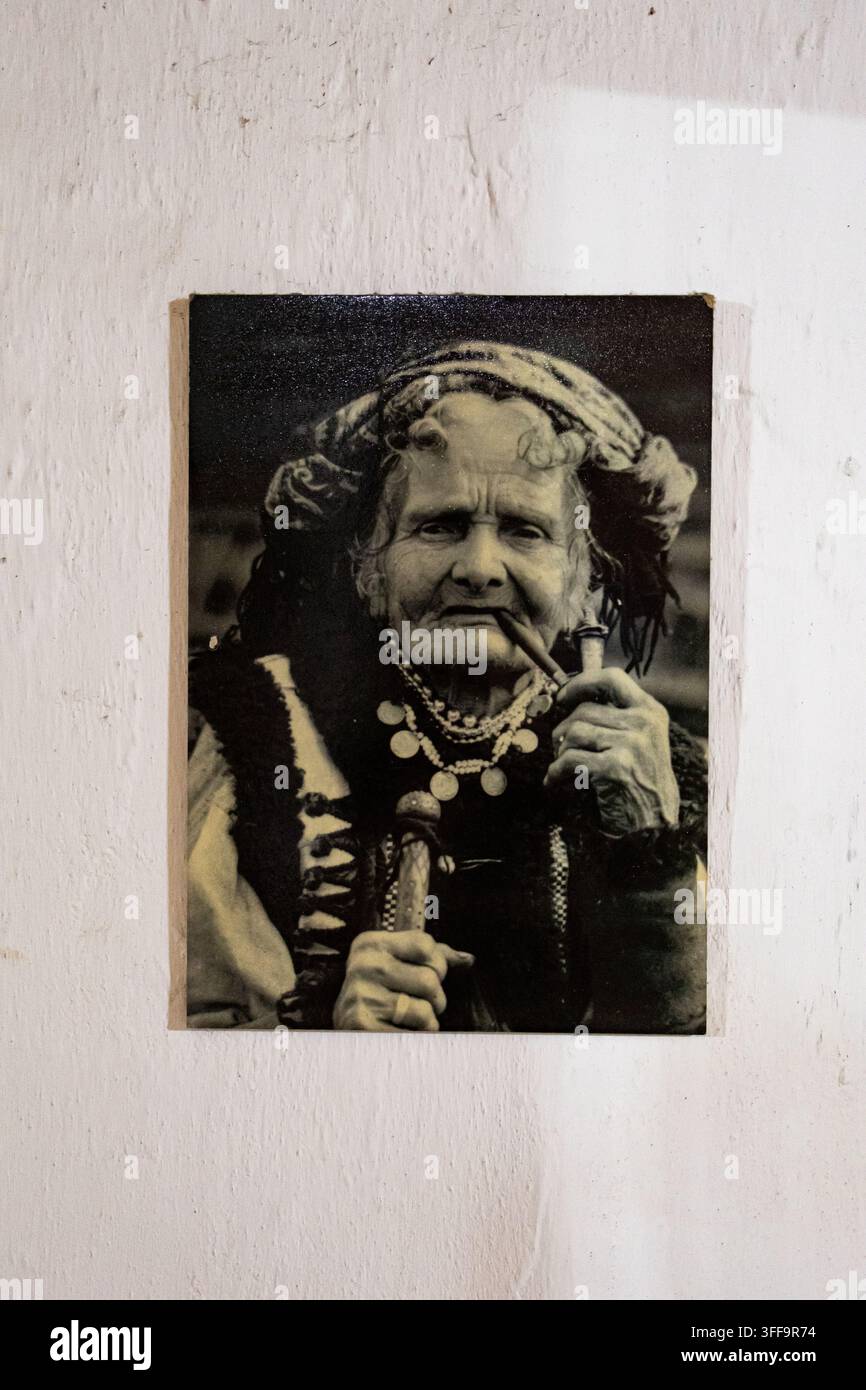 Altes gerahmtes Porträt einer Frau in regionaler Tracht, die eine Pfeife raucht, hängt an einer rauen weißen Wand. Der abgenutzte Druck erinnert an widerstandsfähige Lebensweisen und folkloristische Identität und t Stockfoto