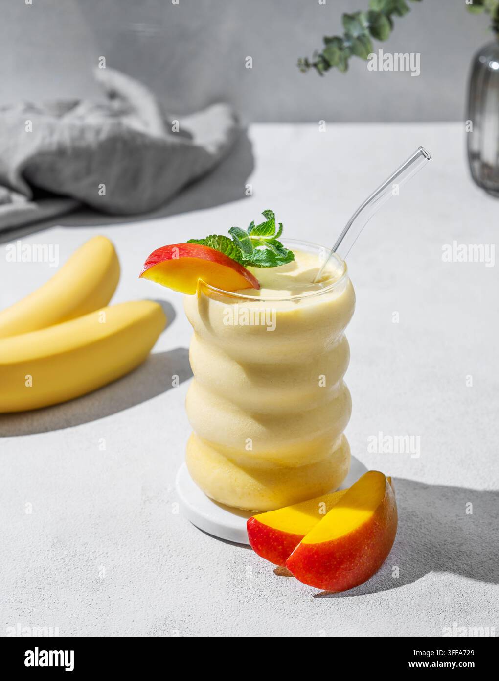 Gefrorener Smoothie mit Mango und Banane in einem Glas auf einem hellen Hintergrund mit tropischen Obstscheiben. Gesundes Essen und Detox-Diät-Konzept. Stockfoto
