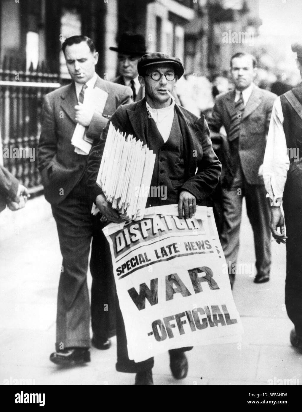 Zeitungsverkäufer in Whitehall, London, am 7. September 1939, der den offiziellen Kriegzustand mit Deutschland verkündet. Er trägt einen Stapel Zeitungen unter einem Arm und zeigt ein großes Plakat mit der Aufschrift „Spezialnachricht: Krieg – Offiziell“. Passanten in Anzügen und Hüten gehen die Straße entlang, während der Verkäufer die Eilmeldung verkündet, und fangen die Dringlichkeit und Atmosphäre der ersten Tage Großbritanniens im Zweiten Weltkrieg ein. Stockfoto