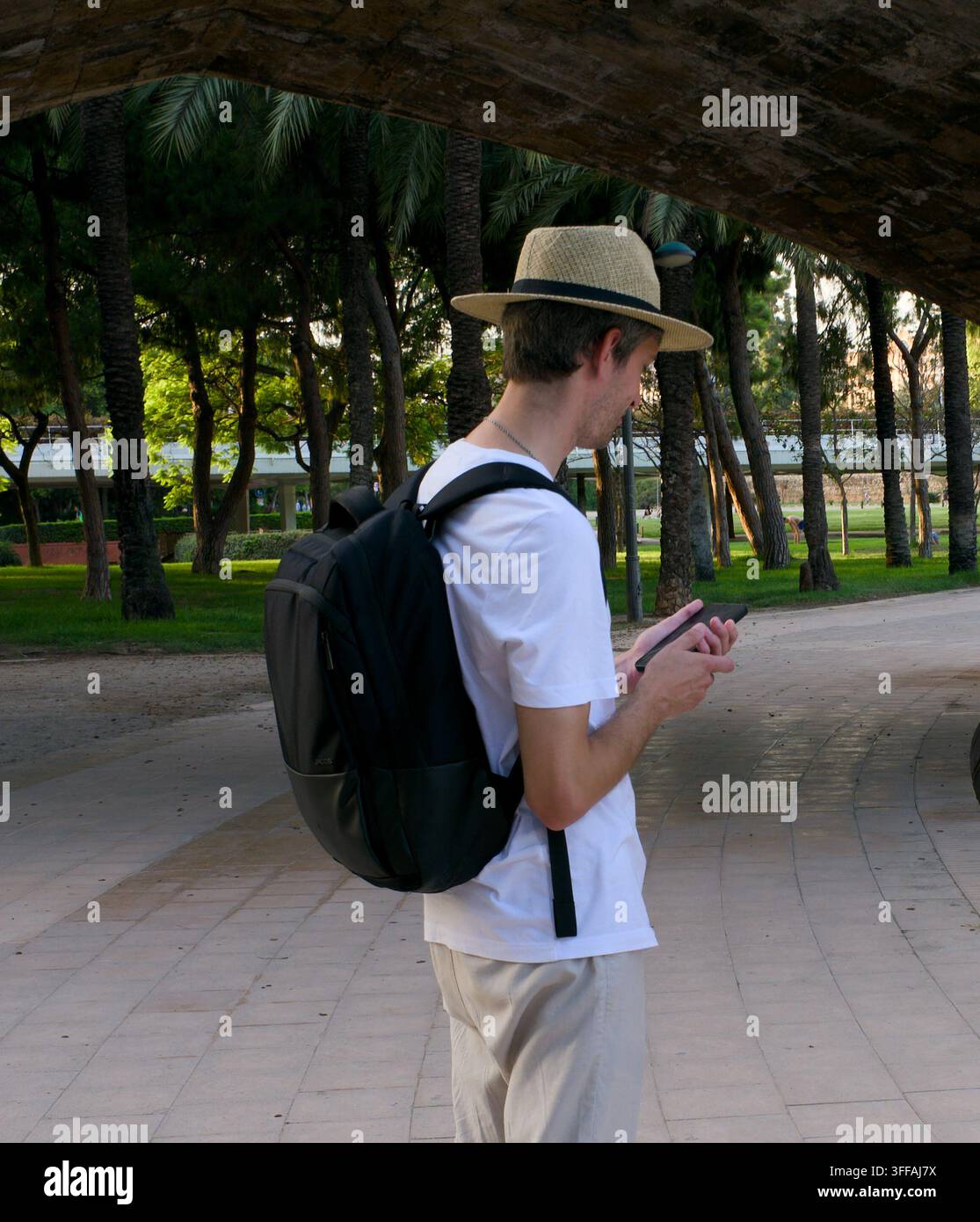 Ein Mann mit Strohhut und Rucksack steht unter einem Brückenbogen, vertieft in sein Smartphone, mit einem üppigen Stadtpark und Palmen im Hintergrund Stockfoto