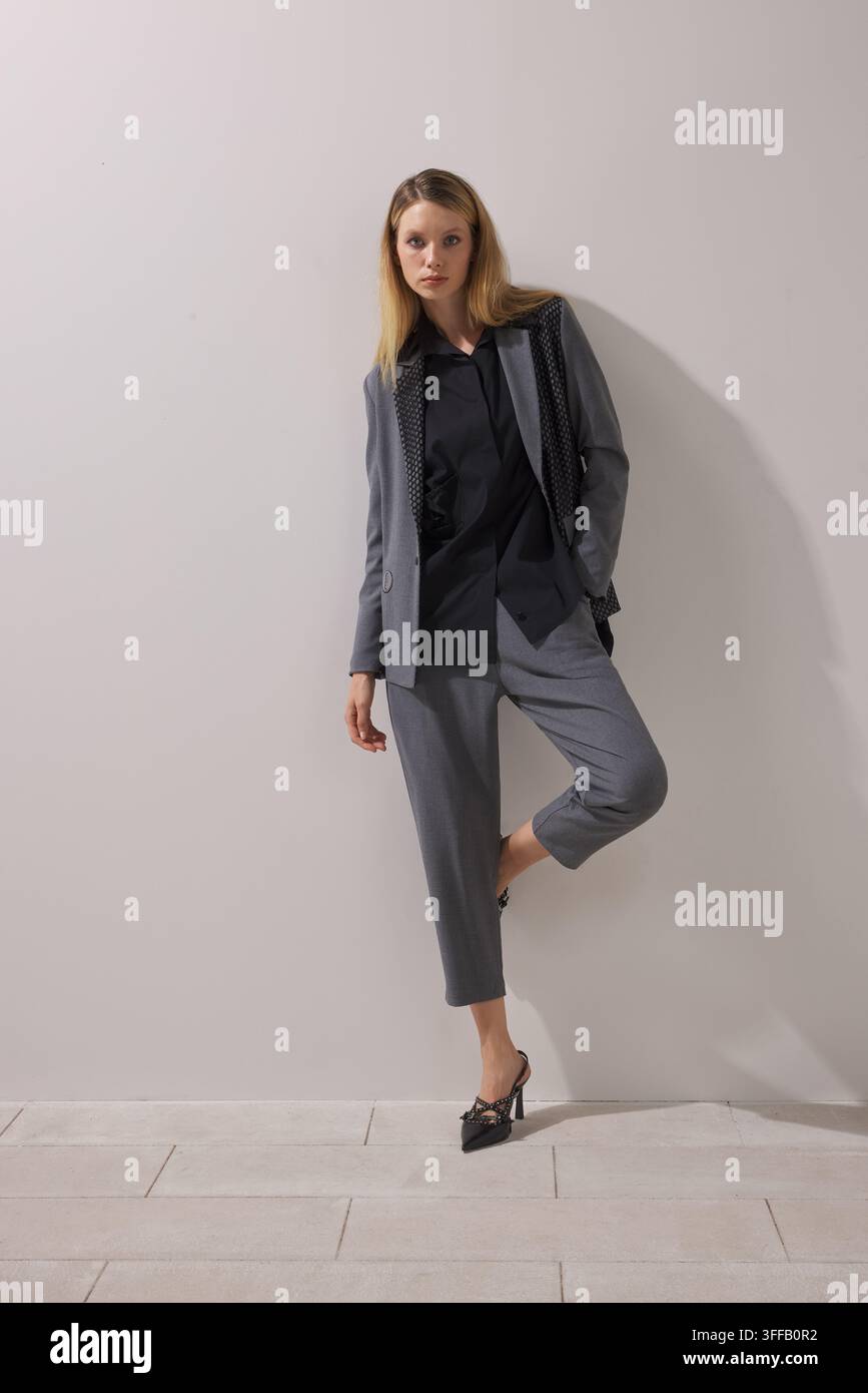 Junge blonde Frau trägt einen grauen oversized Blazer über einem schwarzen Hoodie mit grauen, cropped Tapered-Hosen und schwarzen High Heels. Stockfoto