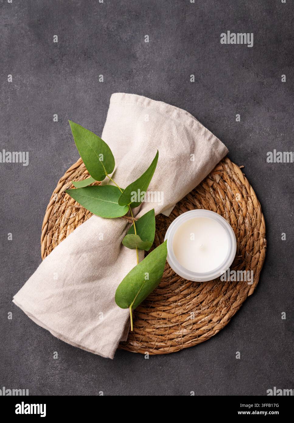 Weiße Kerze im Glas, Eukalyptusblätter und beiger Stoff auf Weidenmatte auf dunklem Betonhintergrund. Natürliches Spa-Konzept. Oberansicht, Kopierraum. Stockfoto