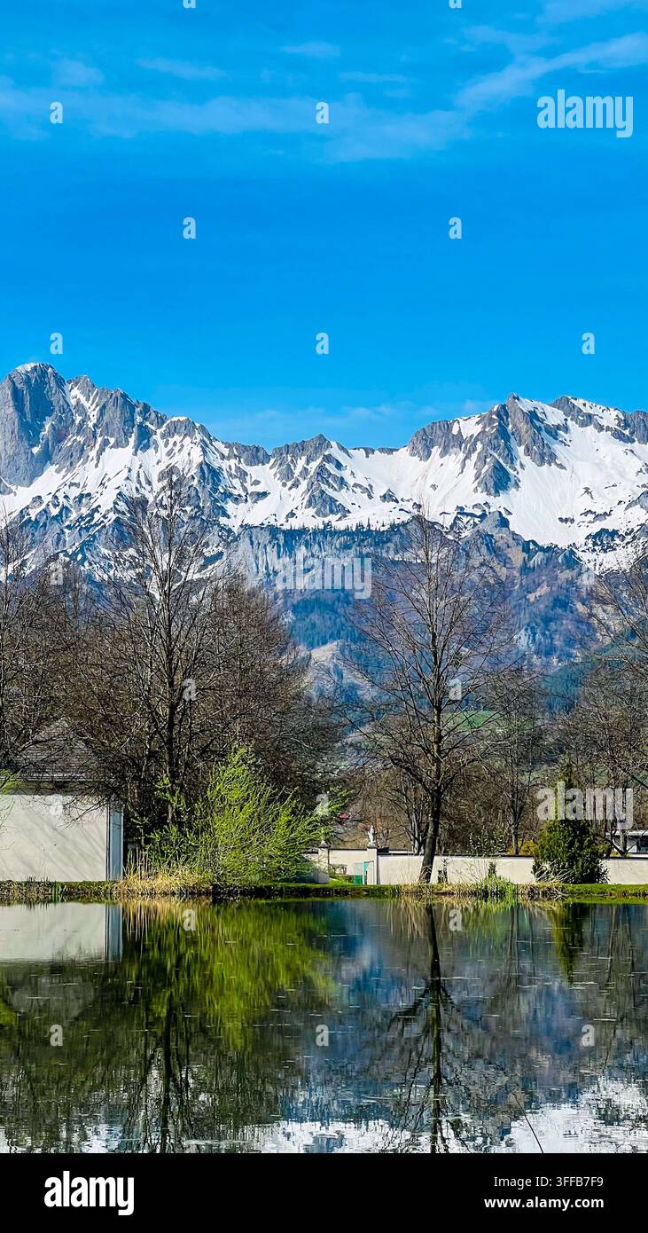 Schneebedeckte Berge erheben sich hinter einem Teich, der Bäume und Himmel widerspiegelt, mit Picea abies und Betula pendula. Konzept der alpinen Natur, des Friedens und des saisonalen Wandels. Stockfoto