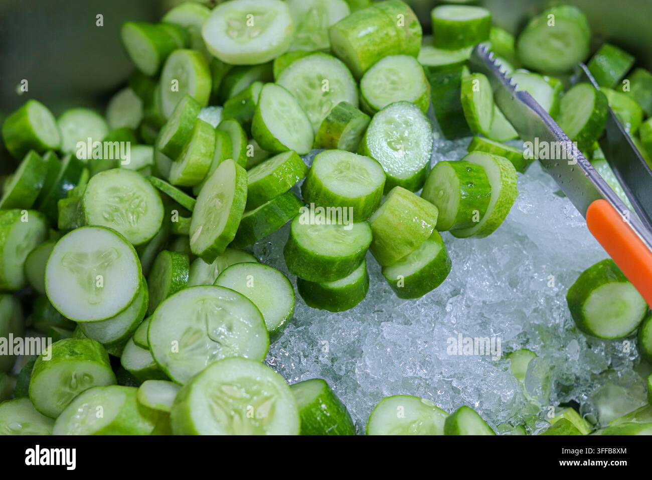 Geschnittene frische grüne Gurken, gekühlt auf zerkleinertem Eis mit Edelstahl-Servierzangen an einem kalten Buffet oder Salatbar. Stockfoto