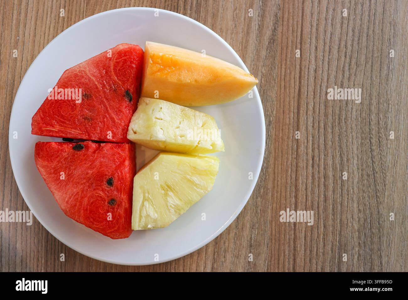 Ansicht von oben: geschnittene Wassermelone, Ananas und Cantaloupe auf einem weißen Teller über einem Holztisch mit freiem Platz auf der rechten Seite. Stockfoto