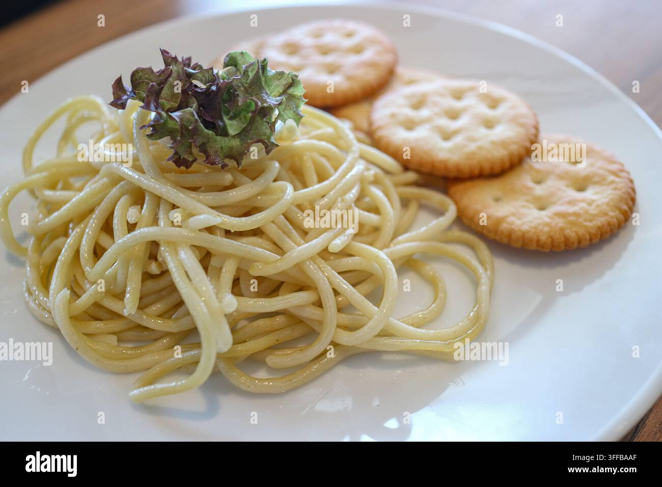 Nahaufnahme von schlicht gekochten Spaghetti, Salzkeksen und rotem Salat auf einem weißen Keramikteller, der auf einem Holztisch ruht, Diätkonzept. Stockfoto