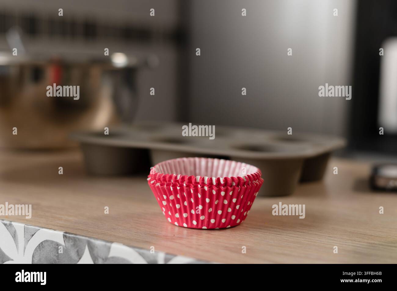 Rosa gepunktete Cupcake-Förmchen, die auf einer Küchentheke gestapelt sind, mit einer Muffinpfanne und einer Edelstahl-Schüssel im Hintergrund. Stockfoto