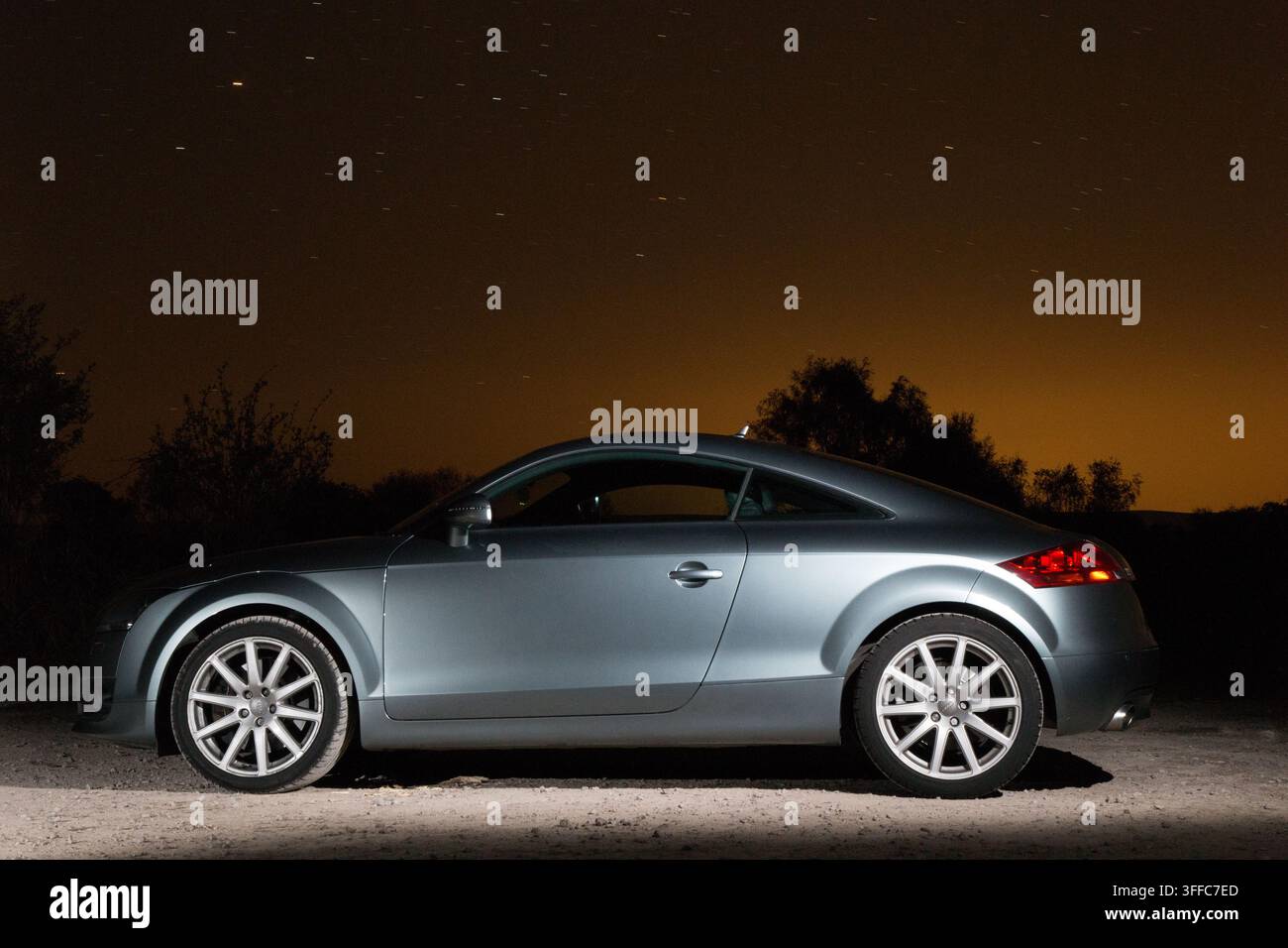 Ein schlankes silbernes Audi TT Coupé, das nachts im Freien geparkt ist, in Seitenansicht unter einem sternenklaren Himmel mit einem warmen bernsteinfarbenen Horizontschein gezeigt. Stockfoto
