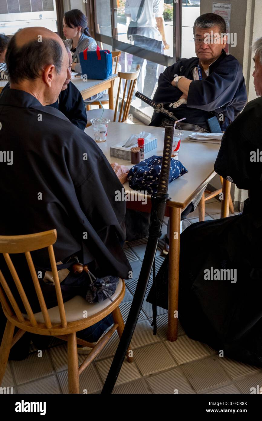 Drei Männer in traditioneller Kleidung mit echten japanischen Katana (刀)-Schwertern, die während einer Kampfkunstausstellung in Kyoto, Japan, in einem Café eine Pause machen. Stockfoto