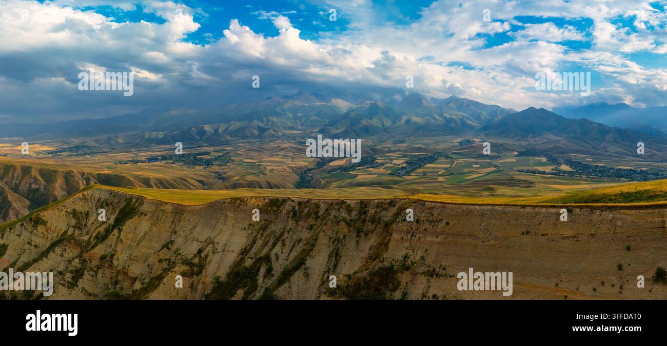 Panoramische Luftaufnahme zeigt eine raue Berglandschaft mit einer steilen Felswand und einem breiten Tal darunter. Sanfte Hügel und ein kleines Dorf sind unter einem dynamischen Himmel mit dramatischen Wolken und Sonnenlicht sichtbar. Stockfoto