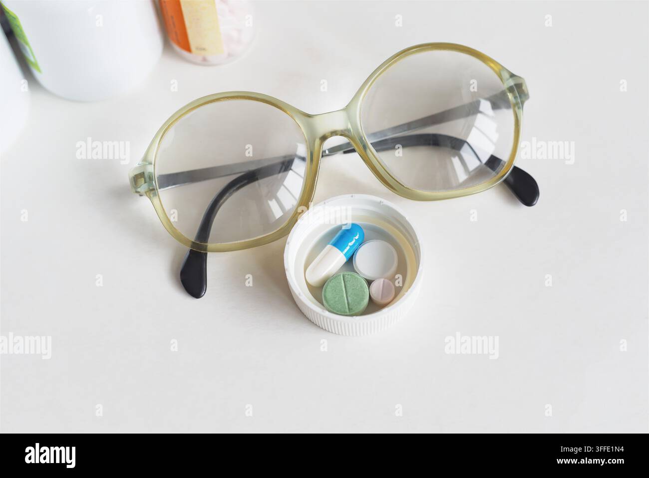 Nahaufnahme von Pillen und Kapseln in einer Dosierkappe neben Lesebrille und Apothekengefäßen auf weißem Hintergrund, für die tägliche Medikamenteinnahme, Senior Stockfoto
