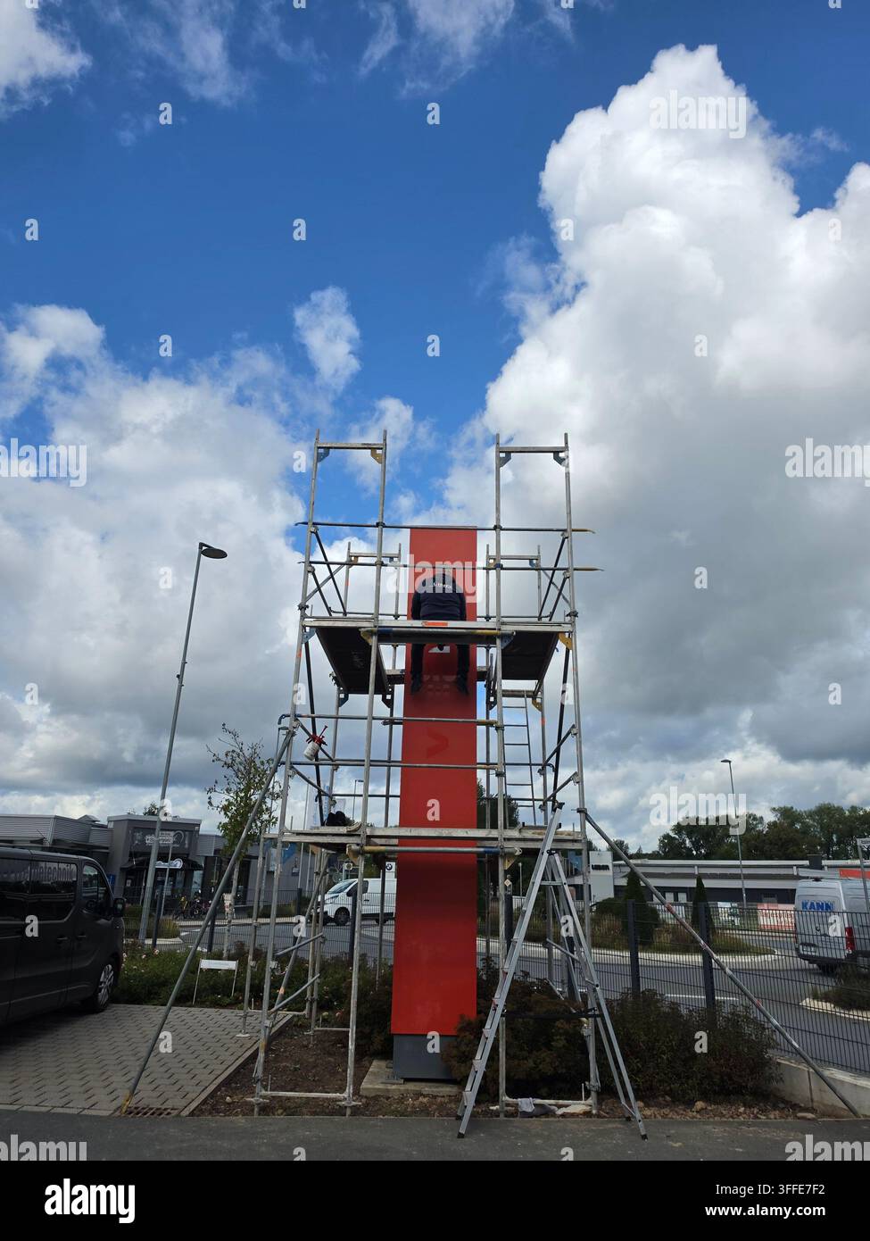 Arbeiter malt rote Struktur auf Metallgerüst während Renovierungsarbeiten in einer deutschen Stadtstraße vor blauem bewölkten Himmel - Smartphone-aufgenommenes Stockfoto